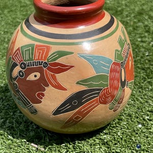 May include: A round, tan ceramic vase with a red rim and decorative bands of green and black. The vase features painted images of stylized figures in red, brown, and black, with green and blue accents. The vase is sitting on green grass.