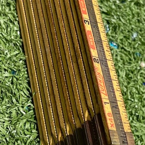 May include: Vintage wooden folding ruler with yellow and brown segments. The ruler has black markings and red text, including the word "Extension". The ruler is on a green surface.