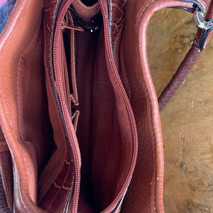 May include: A brown leather handbag with a crocodile-like texture. The bag has a zippered main compartment and multiple interior pockets. The handle is braided, and the hardware is silver-toned. The bag is open, revealing the interior lining.