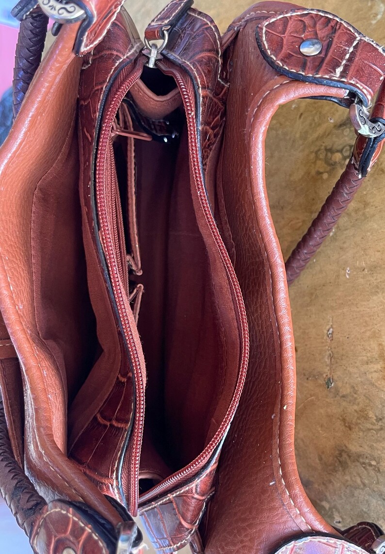 May include: A brown leather handbag with a crocodile-like texture. The bag has a zippered main compartment and multiple interior pockets. The handle is braided, and the hardware is silver-toned. The bag is open, revealing the interior lining.