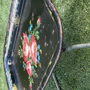 May include: Vintage black metal serving tray with a floral design featuring pink roses and blue accents. The tray is on a folding metal stand, perfect for serving drinks or snacks.