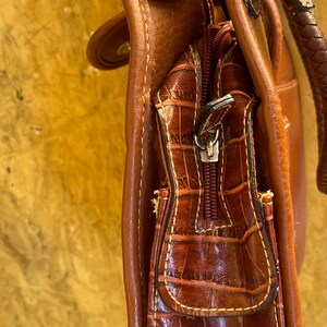 May include: A brown leather handbag with a crocodile-textured front pocket and a zipper closure. The bag has a braided handle and decorative accents. The stitching is visible in a contrasting color.