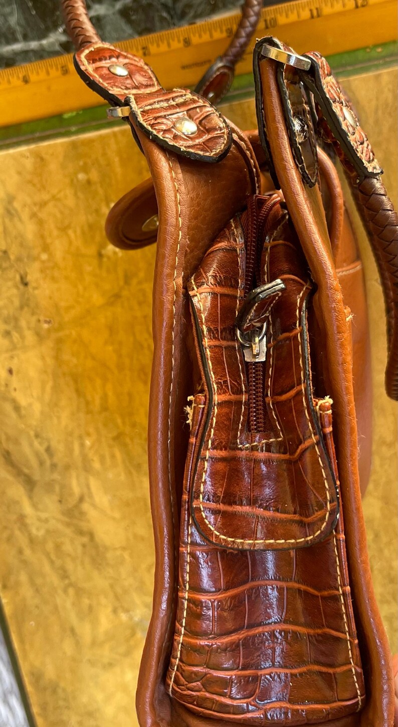 May include: A brown leather handbag with a crocodile-textured front pocket and a zipper closure. The bag has a braided handle and decorative accents. The stitching is visible in a contrasting color.