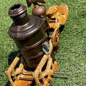 May include: A vintage, cast-iron cannon model with a brown finish. The cannon features an eagle emblem and is mounted on a weathered, tan carriage with wheels. The cannon is set against a green background.