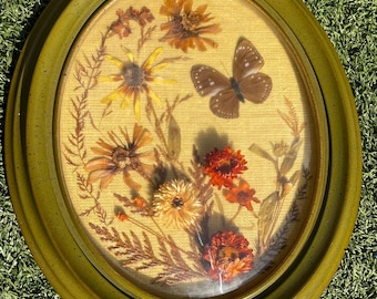 May include: Oval framed artwork featuring dried flowers and a butterfly. The arrangement includes yellow, orange, and brown flowers, with a brown butterfly. The frame is olive green.