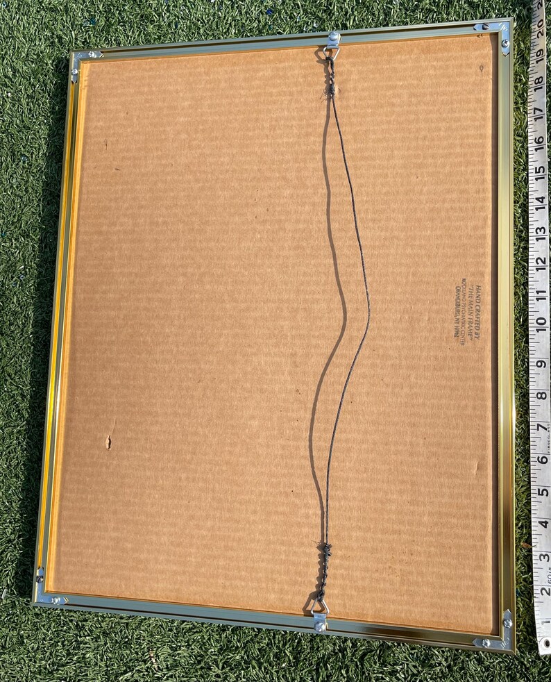May include: Back of a framed artwork with a gold-colored metal frame. The artwork backing is cardboard. A black wire is attached for hanging. The frame has a ruler along the side with inch measurements.