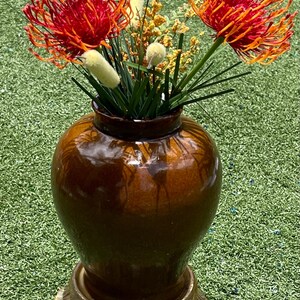 May include: A brown glazed ceramic vase with a decorative base holds a vibrant floral arrangement. The arrangement features red and orange flowers, yellow accents, and green foliage. The vase is approximately 12 inches tall.