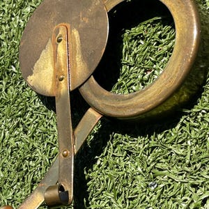 May include: An antique brass pizza cutter with a circular blade and a large ring handle. The blade is connected to the handle by a riveted arm. The cutter is resting on a bed of green artificial grass.