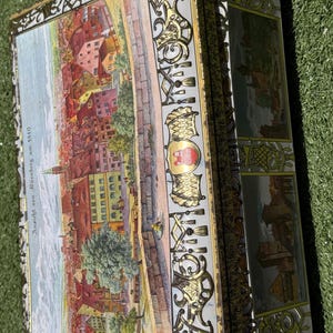 Könnte beinhalten: Rechteckige Vintage-Blechdose mit detaillierter Landschaftsmalerei auf dem Deckel. Die Dose hat einen goldfarbenen, verzierten Rand und den Text "Ansicht von Nürnberg um 1840". Die Seiten der Dose weisen ebenfalls Landschaftsbilder auf.