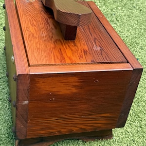 May include: A rectangular wooden shoe shine box with a rich brown stained finish and visible wood grain. The box features a dark brown wooden footrest mounted on the top lid, with metal studs along the side panel and a small silver metal latch.