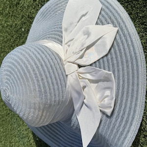 May include: A light blue sun hat with a wide brim and a large white bow. The hat has a textured, ribbed design and a satin ribbon. The bow is made of a silky material.