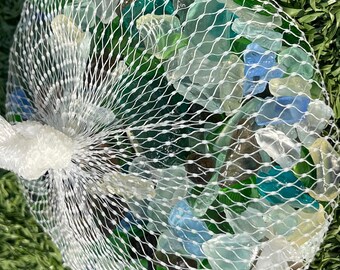May include: A mesh bag filled with assorted sea glass in shades of green, blue, and clear. The bag is tied with a white cord and sits on a bed of green grass. The sea glass appears smooth and tumbled.