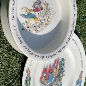May include: Two white ceramic plates featuring illustrations of a rabbit in a blue jacket. The top plate shows the rabbit running from a person with a rake, with text reading: But round the end of a cucumber frame, whom should he meet but Mr. McGregor! he jumped up and ran after Peter, waving a rake and calling out, Stop thief!