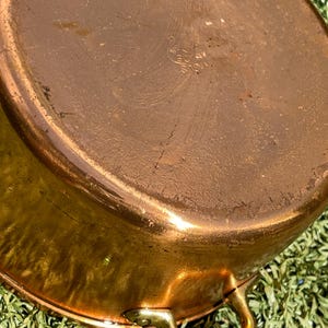 May include: A copper-colored, oval-shaped cooking pot with a brass handle. The pot has a shiny, reflective surface and appears to be vintage. The pot is sitting on a bed of green grass.