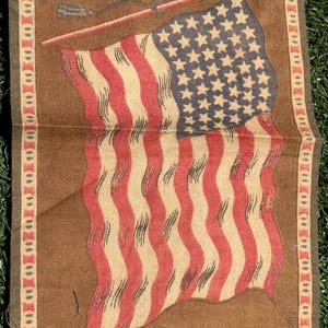 May include: A vintage-style American flag design on a brown fabric. The flag features red and white stripes with a blue field of stars. The border has a decorative pattern with blue accents. The flag is likely a decorative item.