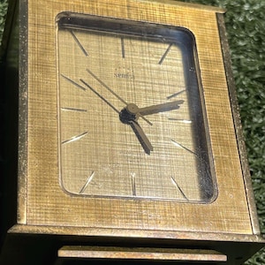 May include: A vintage gold-toned Semca clock with a rectangular face. The clock features a textured gold face with black hands and hour markers. The clock's frame has a textured design. The word "Semca" is printed on the clock face.
