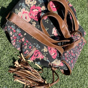 May include: Black floral print tote bag with brown leather straps and trim. The bag features a pattern of pink and red roses with green leaves. A brown leather tassel hangs from the side.