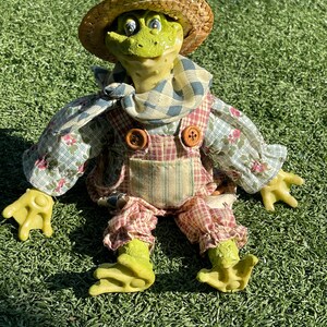 May include: A whimsical frog figurine wearing a straw hat, overalls, and a patterned shirt. The frog has green skin, yellow hands and feet, and is sitting on green grass. The overalls have large wooden buttons.