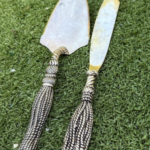 May include: A cake server and butter knife set with silver blades and ornate, tassel-like handles. The blades have a silver finish, and the handles are a metallic silver color. The set is displayed on a bed of green grass.