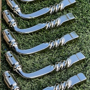 May include: A collection of eight polished chrome door handles. Each handle has a curved design with a cylindrical end and a twisted, ridged section leading to a rectangular base. The handles are arranged on a bed of green grass.