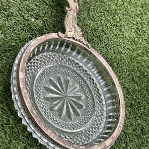 May include: A vintage glass serving dish with a silver-toned metal handle and rim. The clear glass features a diamond-cut pattern and a starburst design in the center. The handle has an ornate, decorative design.