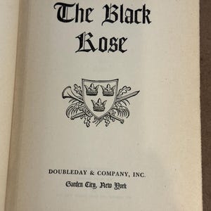 May include: Open book page with the title "The Black Rose" in ornate black lettering. The author's name, Thomas B. Costain, is above the title. A black and white emblem with three crowns is below the title. The publisher's name, Doubleday & Company, Inc., and the location, Garden City, New York, are at the bottom.
