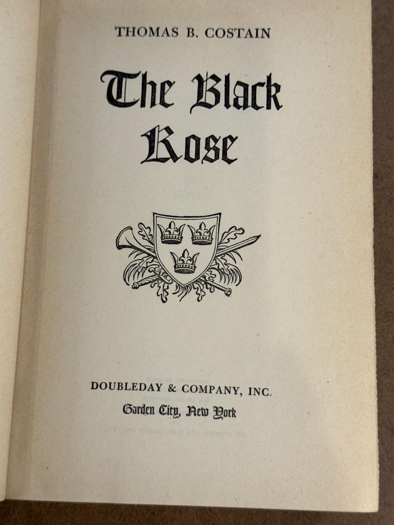 May include: Open book page with the title "The Black Rose" in ornate black lettering. The author's name, Thomas B. Costain, is above the title. A black and white emblem with three crowns is below the title. The publisher's name, Doubleday & Company, Inc., and the location, Garden City, New York, are at the bottom.