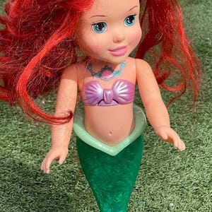 Pode incluir: Boneca de sereia com cabelo vermelho brilhante, olhos azuis e um top rosa em forma de la&ccedil;o. A sereia tem uma cauda verde e uma gola verde clara. A boneca est&aacute; sobre uma superf&iacute;cie verde.
