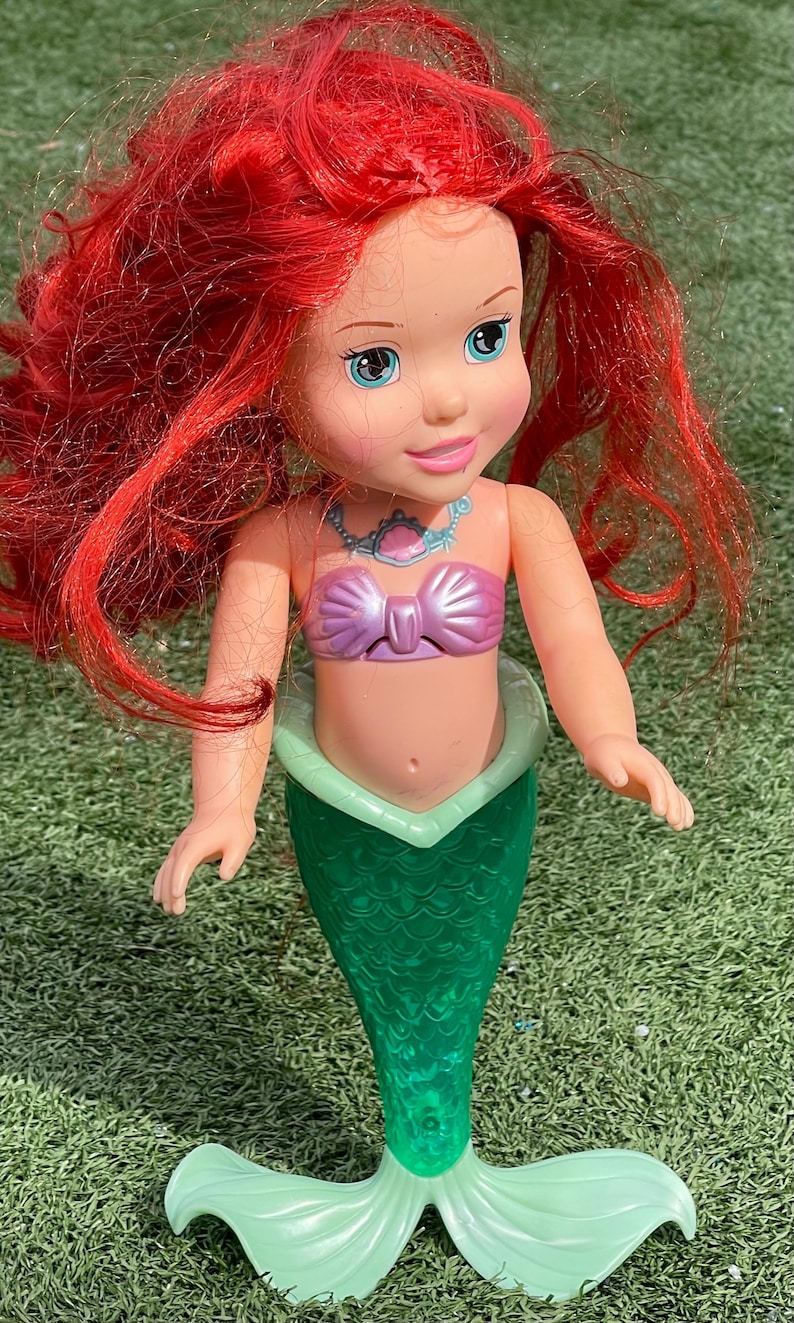 Pode incluir: Boneca de sereia com cabelo vermelho brilhante, olhos azuis e um top rosa em forma de la&ccedil;o. A sereia tem uma cauda verde e uma gola verde clara. A boneca est&aacute; sobre uma superf&iacute;cie verde.