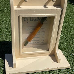 May include: A cream-colored wooden display with a framed section holding a rolling pin and a recipe card. The recipe card has handwritten text. Two small knobs are at the top. The display is on a green surface.