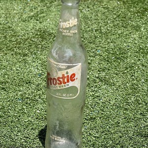 May include: A clear glass Frostie root beer bottle with a white and red label. The label reads "Frostie Root Beer" and "16 fl oz (1 pt)". The bottle also has the text "Money Back Bottle" and "Return for Refund".