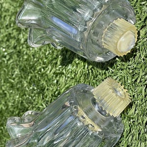May include: Two clear glass candle holders with a starburst design on the top and a yellow-tinted base. The candle holders are sitting on a bed of green grass.
