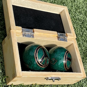 May include: Two green decorative spheres with copper-colored accents and peace symbols, displayed in a light wooden box with a black velvet interior. The box is open, revealing the spheres. The spheres have a marbled appearance.