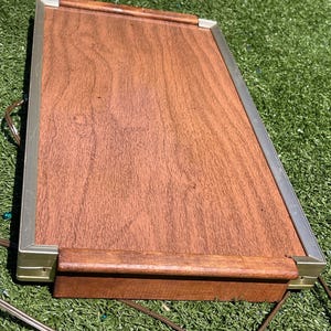 May include: A rectangular, wooden serving tray with a brown finish and a metal frame. The tray has a handle on one side and a power cord. The tray is sitting on a green surface.
