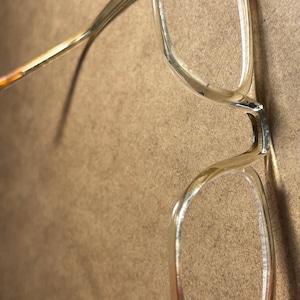 May include: A pair of eyeglasses with a clear, rectangular frame and a gradient of colors from light brown to clear. The temples are thin and metallic, with a similar color gradient. The glasses are set against a neutral background.