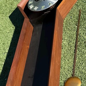 May include: A mid-century modern wooden grandfather clock with a silver-colored clock face and black hands. The clock has a long, rectangular body with a pendulum and a brass-colored weight. The clock is set on a green surface.