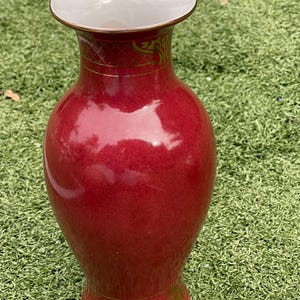 May include: A tall, red ceramic vase with a wide, white interior and a gold rim. The vase features gold decorative patterns around the neck and base. The vase is set against a green background.