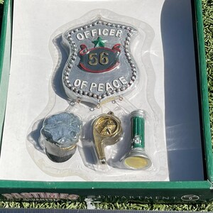 May include: A set of miniature police-themed ornaments, including a badge with "OFFICER OF PEACE 56", a cap, a whistle, and a green flashlight. The ornaments are silver, gold, and green, packaged in a green box.