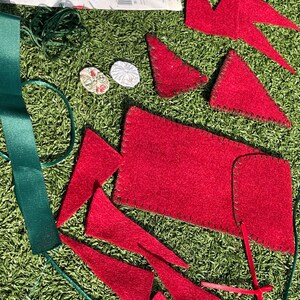 May include: A collection of red felt pieces, including triangles and a rectangle, are scattered on a green surface. Green ribbon and string are also present, along with small decorative buttons and a paper with text. Crafting supplies.