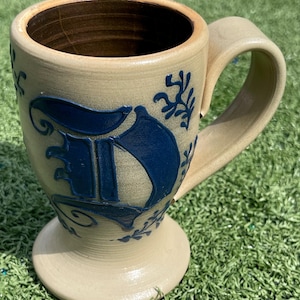 May include: A beige ceramic mug with a large blue letter "F" and floral accents. The mug has a rounded base, a curved handle, and a dark brown interior. The design is reminiscent of traditional pottery.
