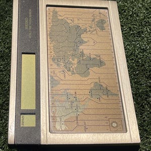 May include: A vintage Seiko World Time Touch Sensor device. The device has a rectangular shape with a silver frame and a world map display. The display shows time zones and cities. The top of the device has the text "SEIKO WORLD TIME TOUCH SENSOR".