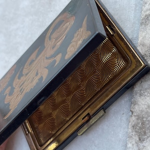 May include: An open, vintage compact case with a black exterior and gold accents. The lid features a gold-colored, stylized design. The interior has a gold-colored, circular pattern. The compact is rectangular and has a clasp.