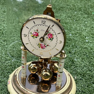 May include: An antique clock with a cream-colored face featuring floral accents and gold hands. The clock has a decorative base with floral patterns and gold accents. A glass dome covers the clock, which is set on a green surface.