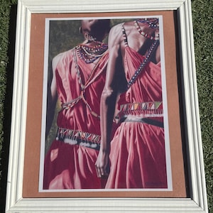 May include: Framed print featuring two figures in traditional attire. The figures wear flowing, coral-colored dresses and beaded necklaces. The print is set in a white frame with a brown matting, creating a striking contrast.