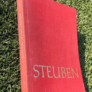 May include: A red book with the word "STEUBEN" in gold lettering on the cover. The spine reads "STEUBEN GLASS 1903-1970". The book is set against a green background.