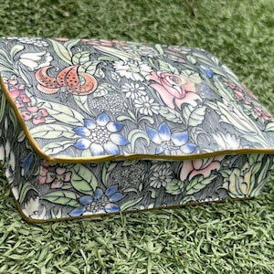 May include: A rectangular jewelry box with a floral pattern. The box features a dark gray background with colorful flowers, including pink roses, blue blossoms, and orange lilies. The edges are trimmed with gold.