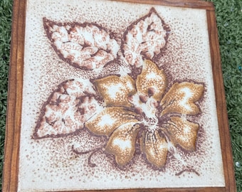 May include: Square decorative tile with a floral design. The tile features a brown and tan flower and leaves on a speckled background, set within a wooden frame. The floral design is detailed and textured, adding visual interest to the piece.