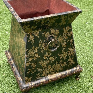 May include: A decorative, square planter with a dark green floral pattern and bronze hardware. The planter has a dark brown interior and sits on four small, decorative feet. A bronze ring handle is attached to the side.