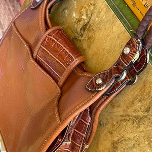 May include: A brown leather handbag with a braided handle and decorative crocodile-textured accents. The bag features a front pocket and a zippered compartment. Silver-colored hardware and small decorative gems are visible on the straps.
