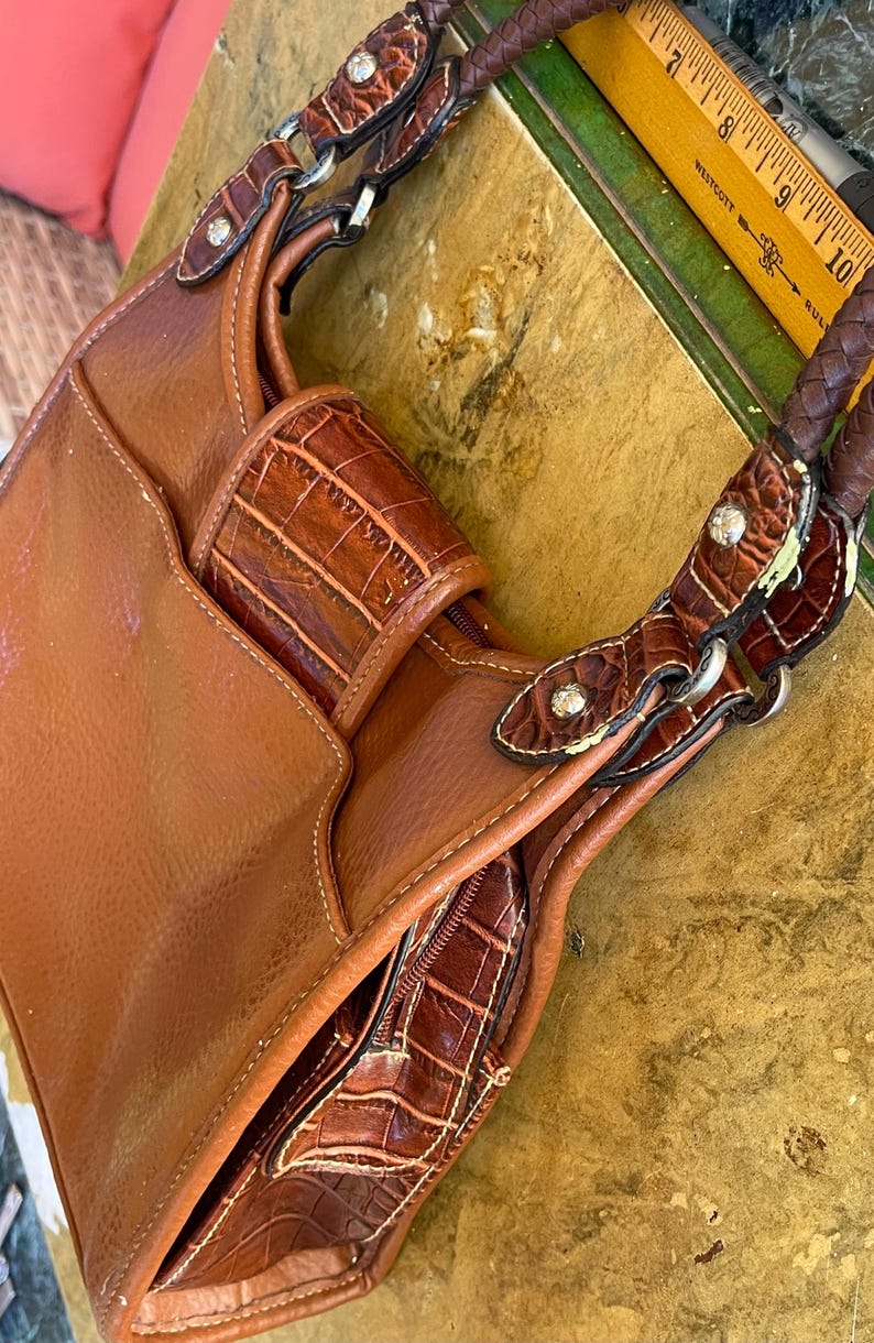 May include: A brown leather handbag with a braided handle and decorative crocodile-textured accents. The bag features a front pocket and a zippered compartment. Silver-colored hardware and small decorative gems are visible on the straps.
